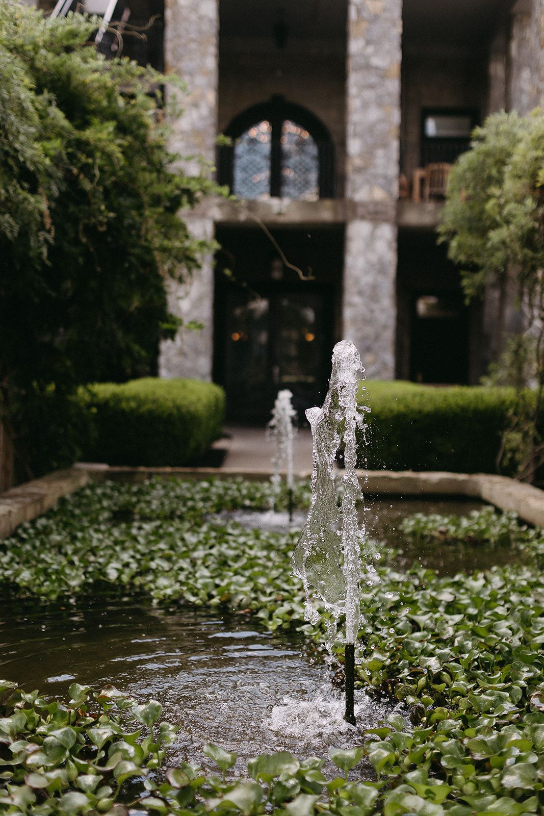 Small courtyard fountain with water jets, green ivy, and stone arches in the background