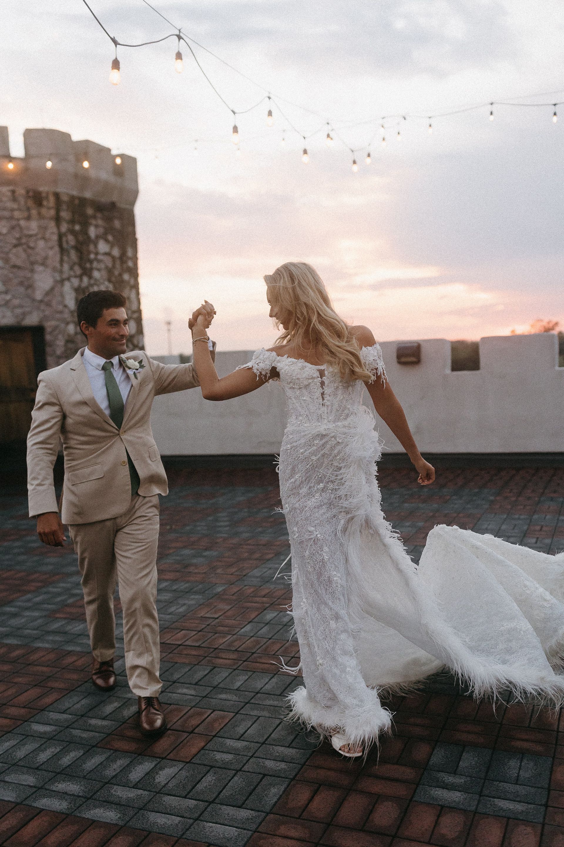 Bride and groom celebrate on a rooftop at sunset. She wears a white gown, he a tan suit, both smiling.