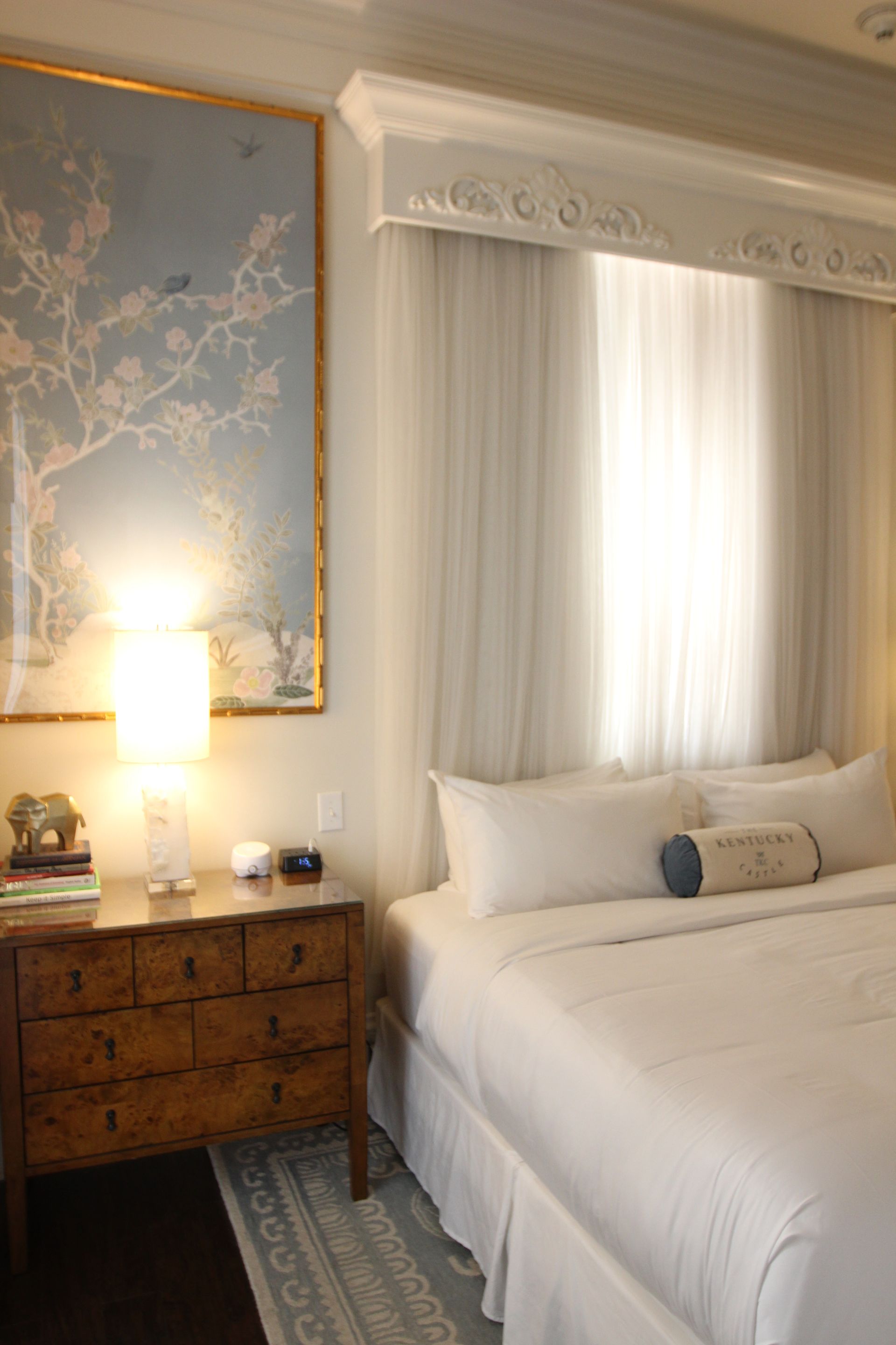 Bedroom with a bed, dresser, large art piece, and sheer curtains. Soft whites and a pale blue color palette.