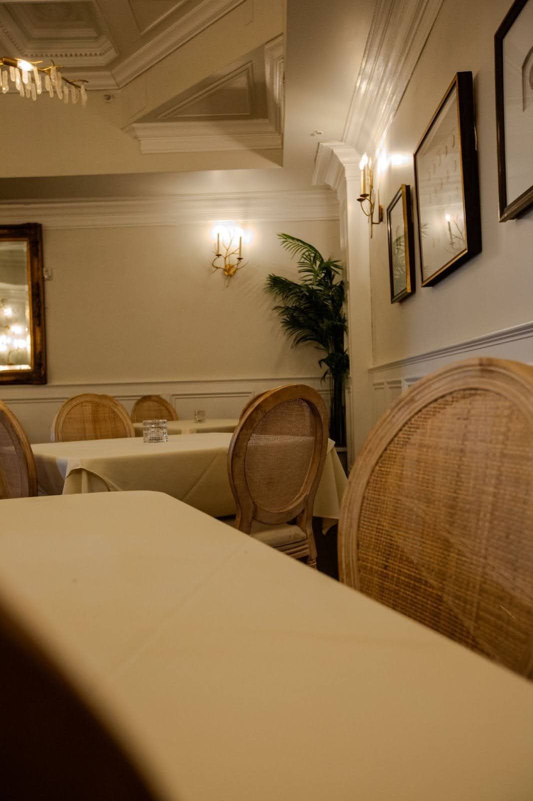 Restaurant dining room with rattan chairs, white tablecloths, sconces, and framed art.