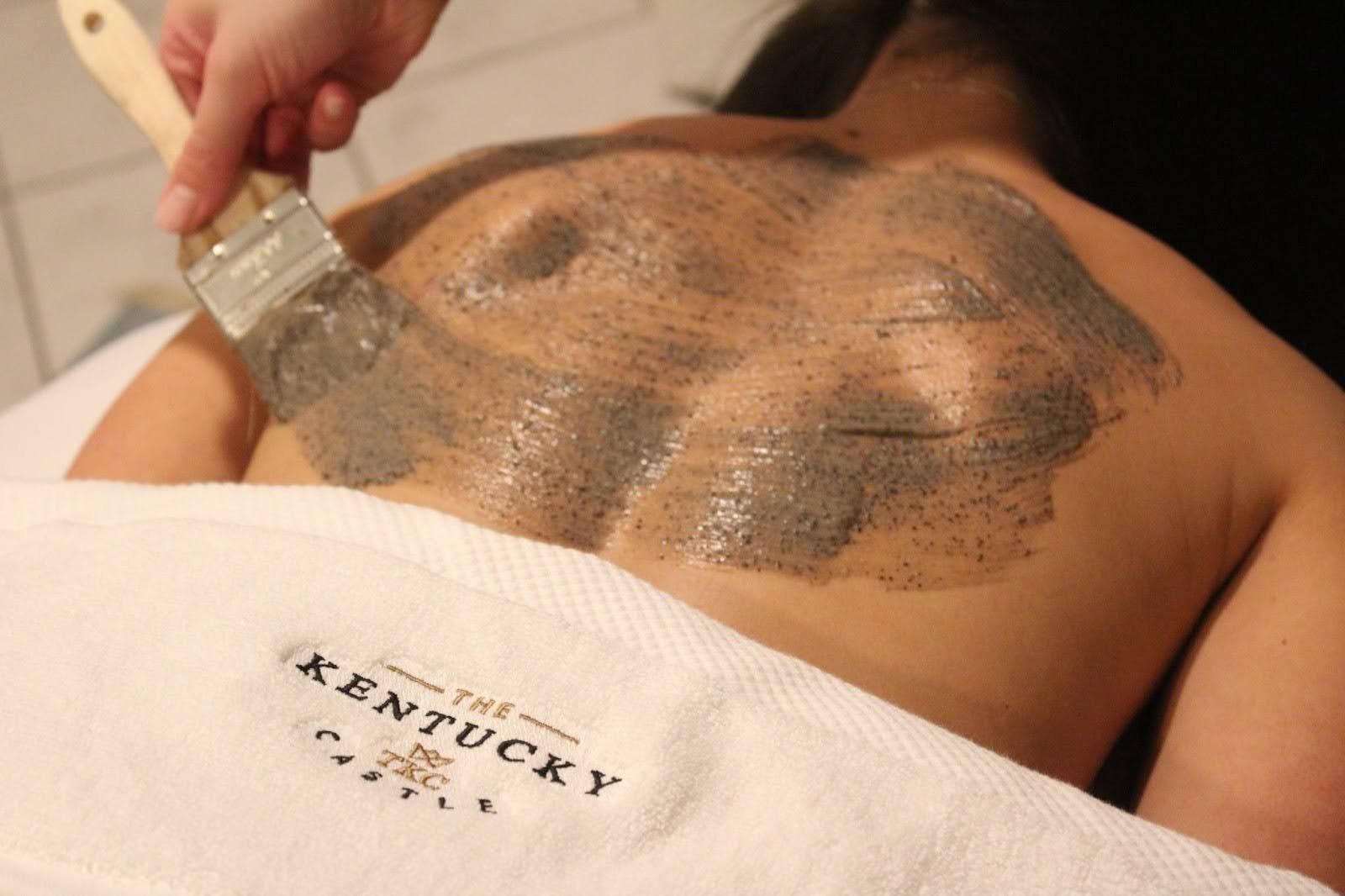 Person receiving back treatment with a brush applying a mud exfoliator at a spa.