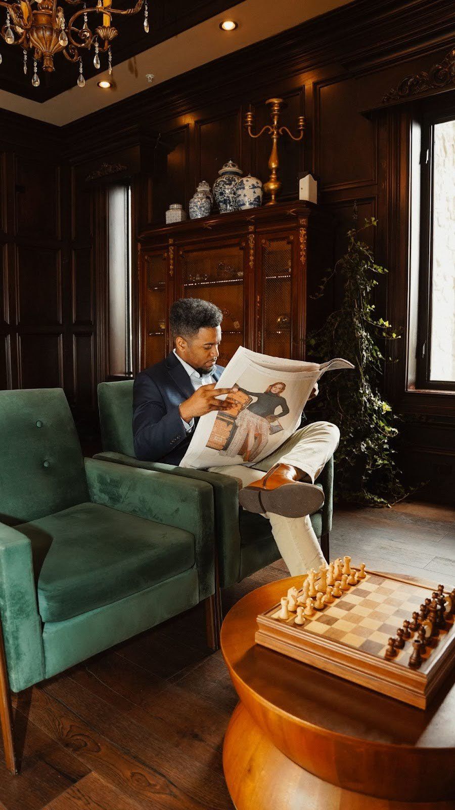 Man in suit reads newspaper in a green armchair, next to a chessboard, in a room with dark wood paneling.