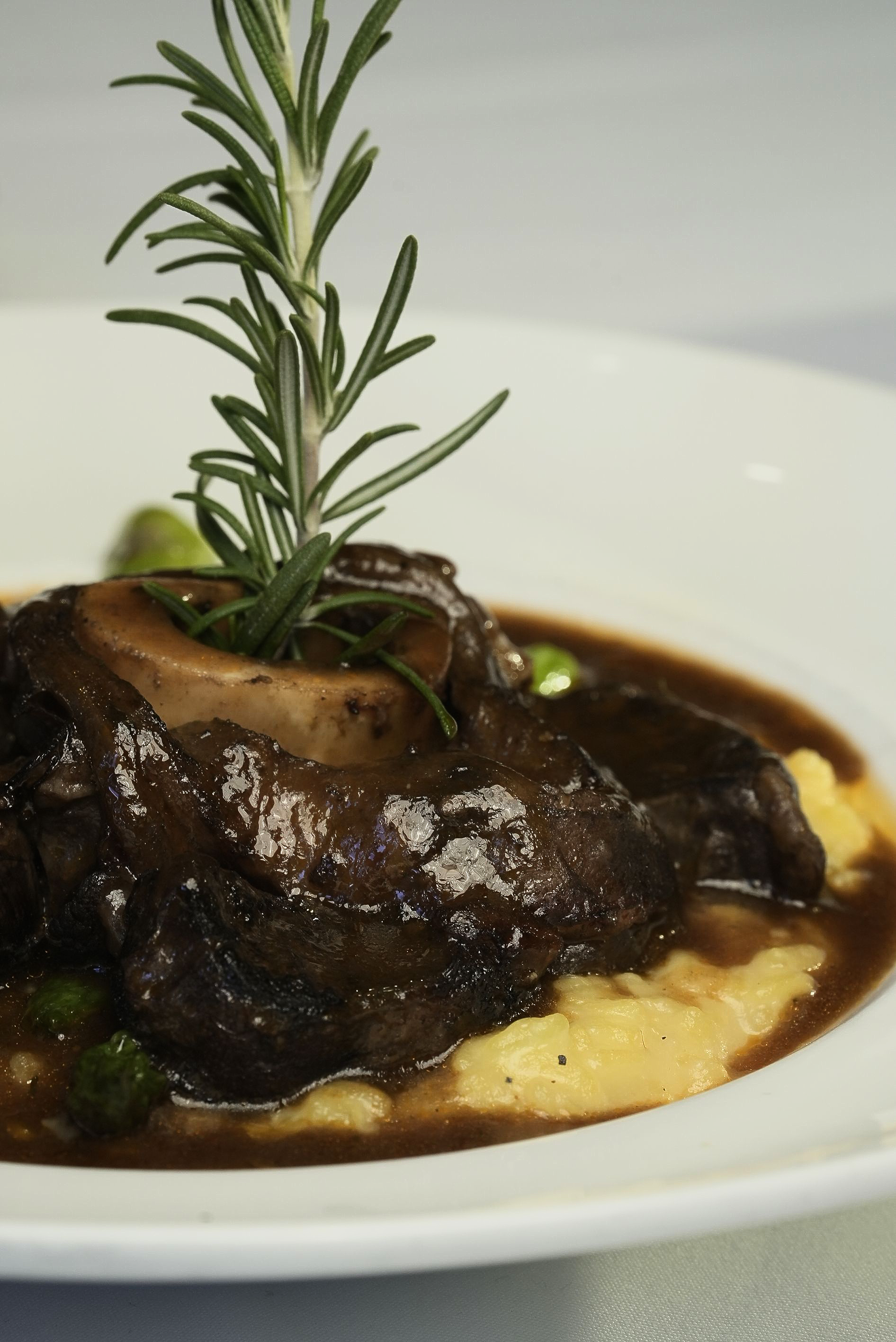 Osso buco with polenta and rosemary sprig garnish in a white bowl.
