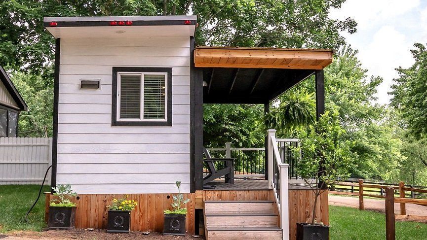 Tiny white house with porch, black trim, small steps, surrounded by greenery.