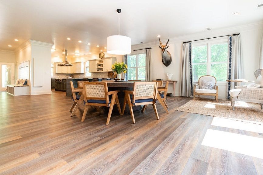 Spacious dining area with wooden floor, table, chairs, and a mounted deer head. Kitchen in the background.