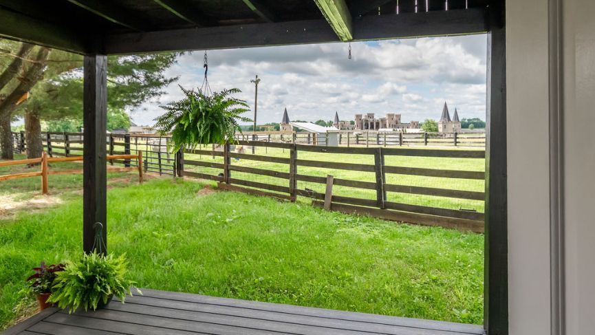 View from a porch with hanging ferns, overlooking a grassy yard with a wooden fence and a farm in the distance.