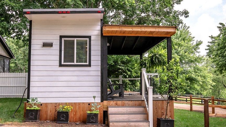 White tiny house with black trim, small porch, and a window with blinds.