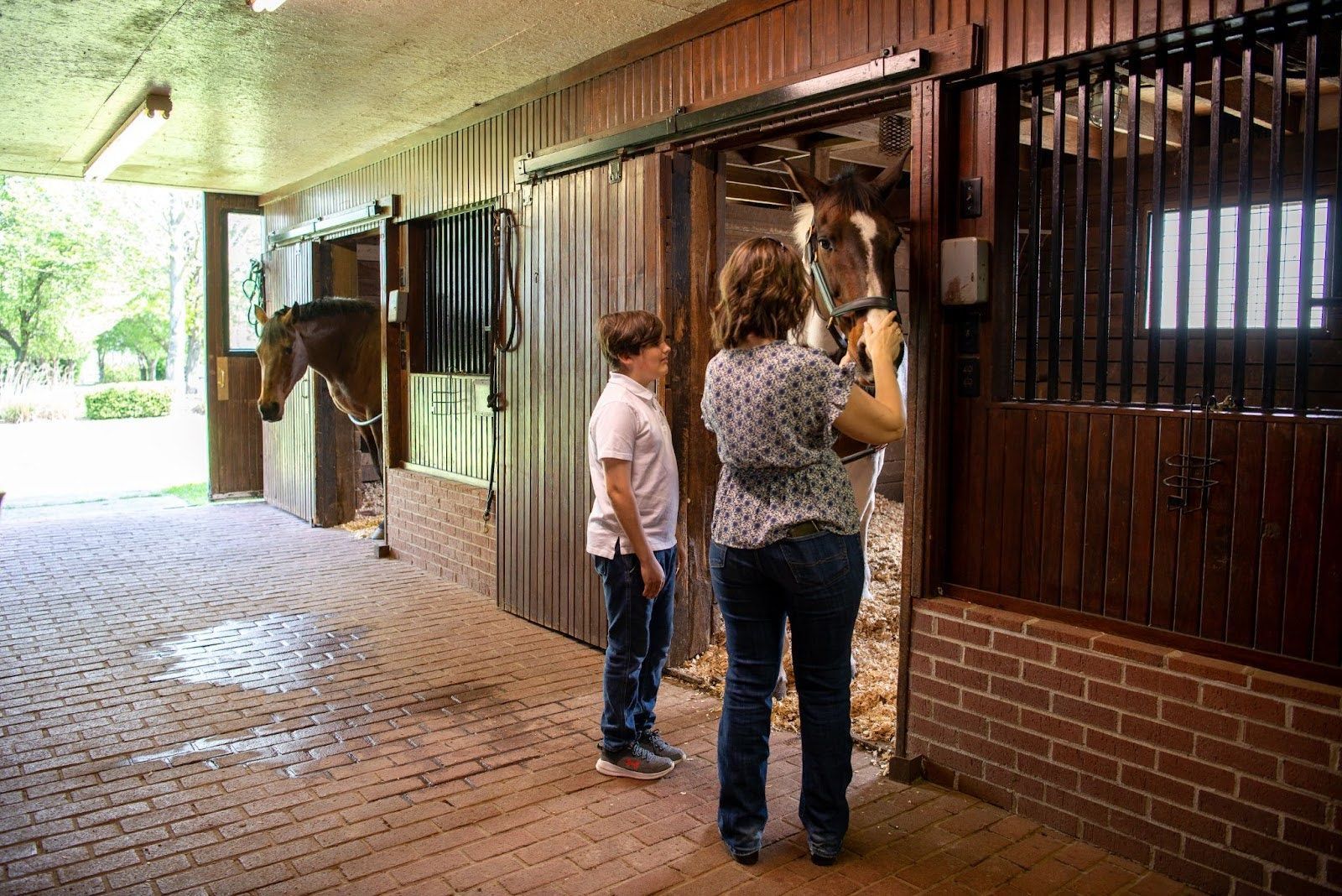 Woman and child interacting with horse in a stable. Brown wood and brick interior.