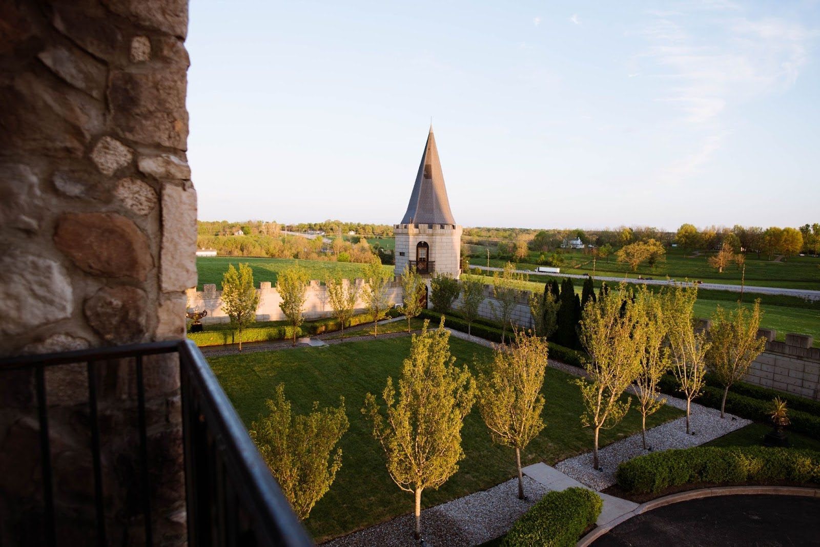 View from a stone castle balcony of a manicured green lawn with trees and a turret in the background.