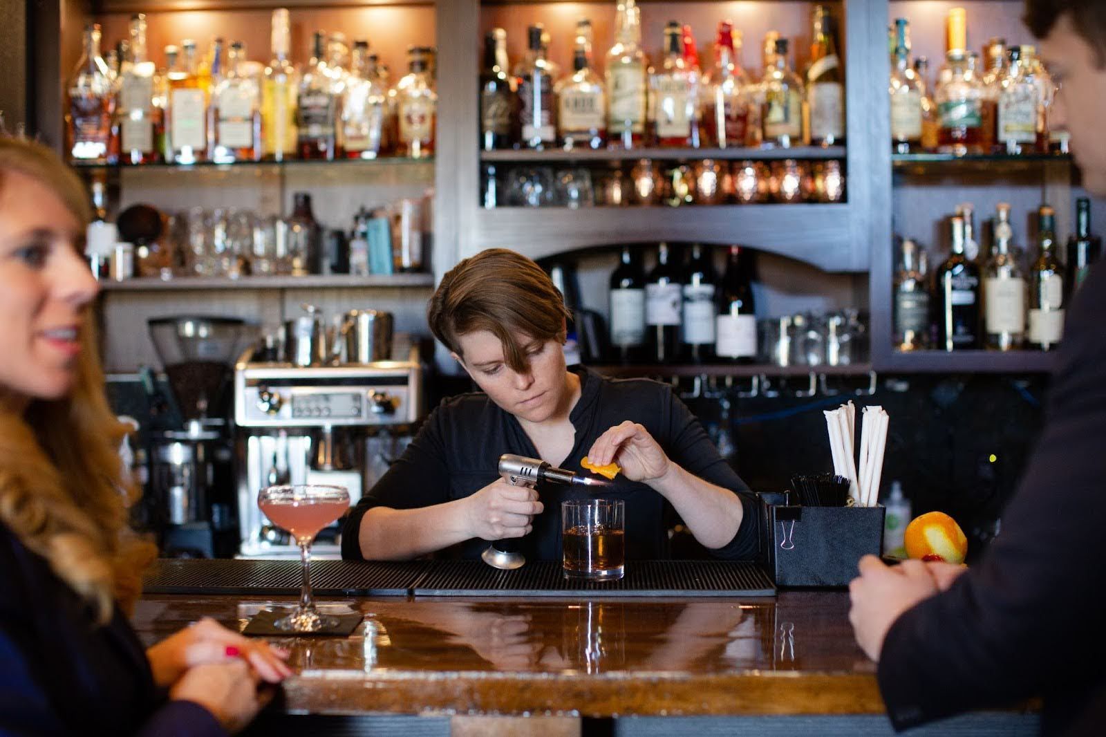 Bartender torches an orange peel over a drink, preparing a cocktail at a bar. Customers nearby.