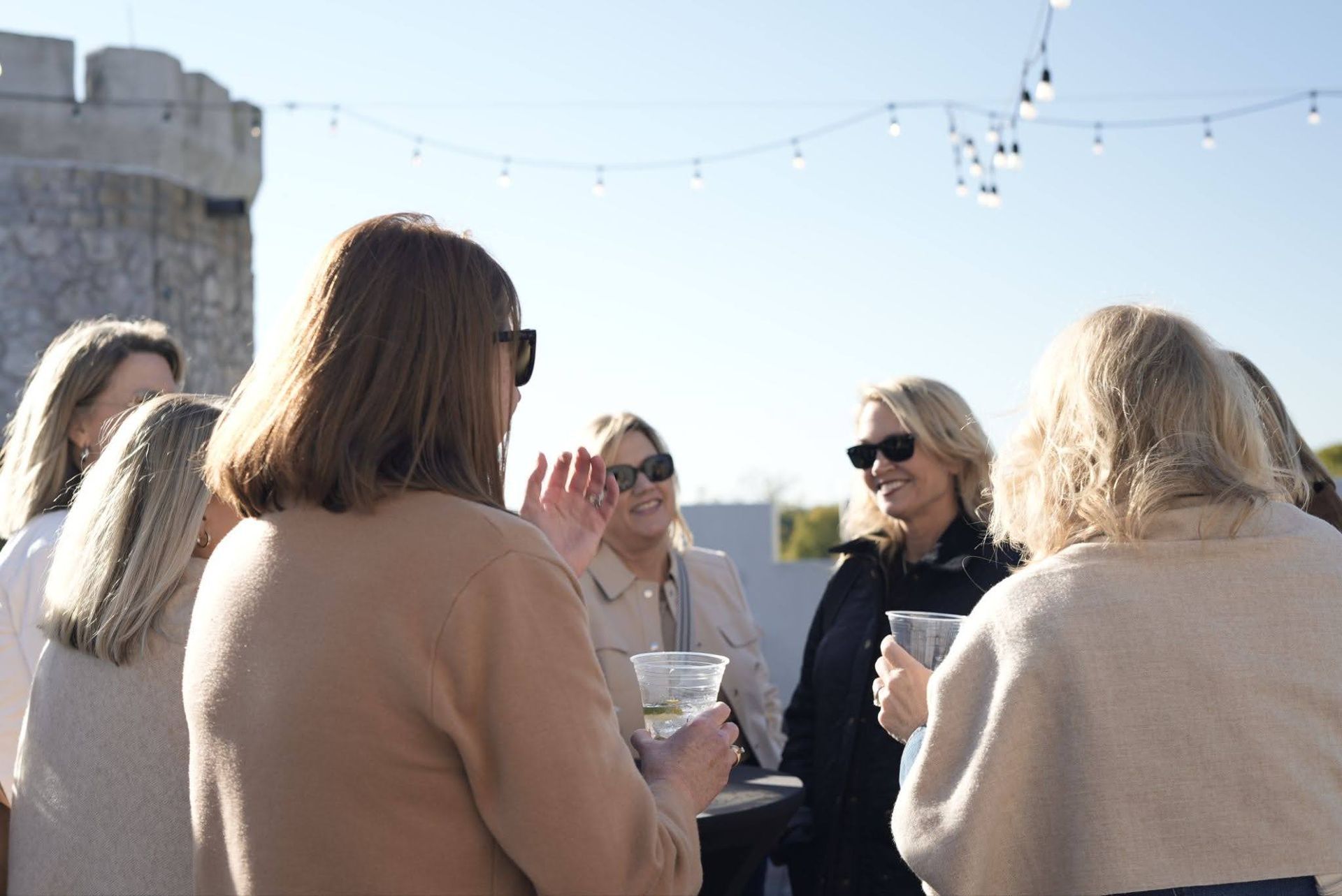 People chatting outdoors in sunlight near a stone wall, some holding drinks and wearing sunglasses.