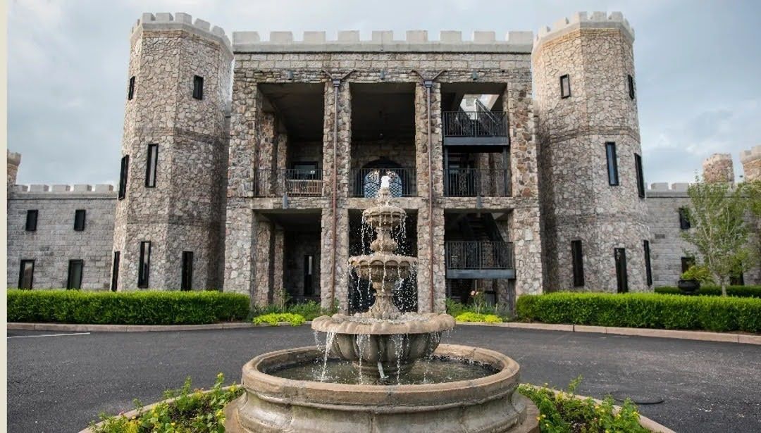 Stone castle-like building with twin towers behind a central fountain and manicured hedges