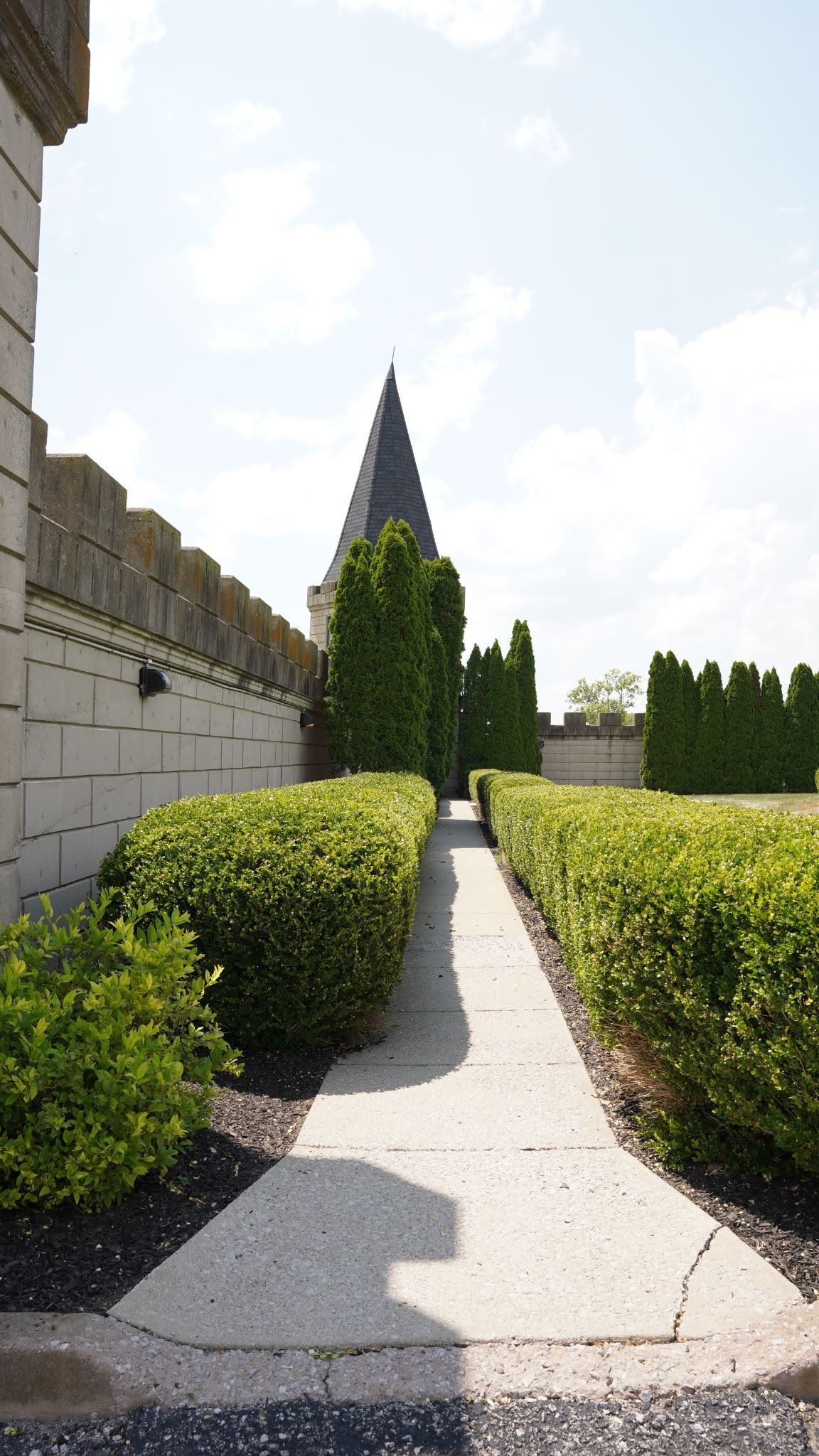 Sunlit garden path between trimmed hedges leading toward a tall pointed tower