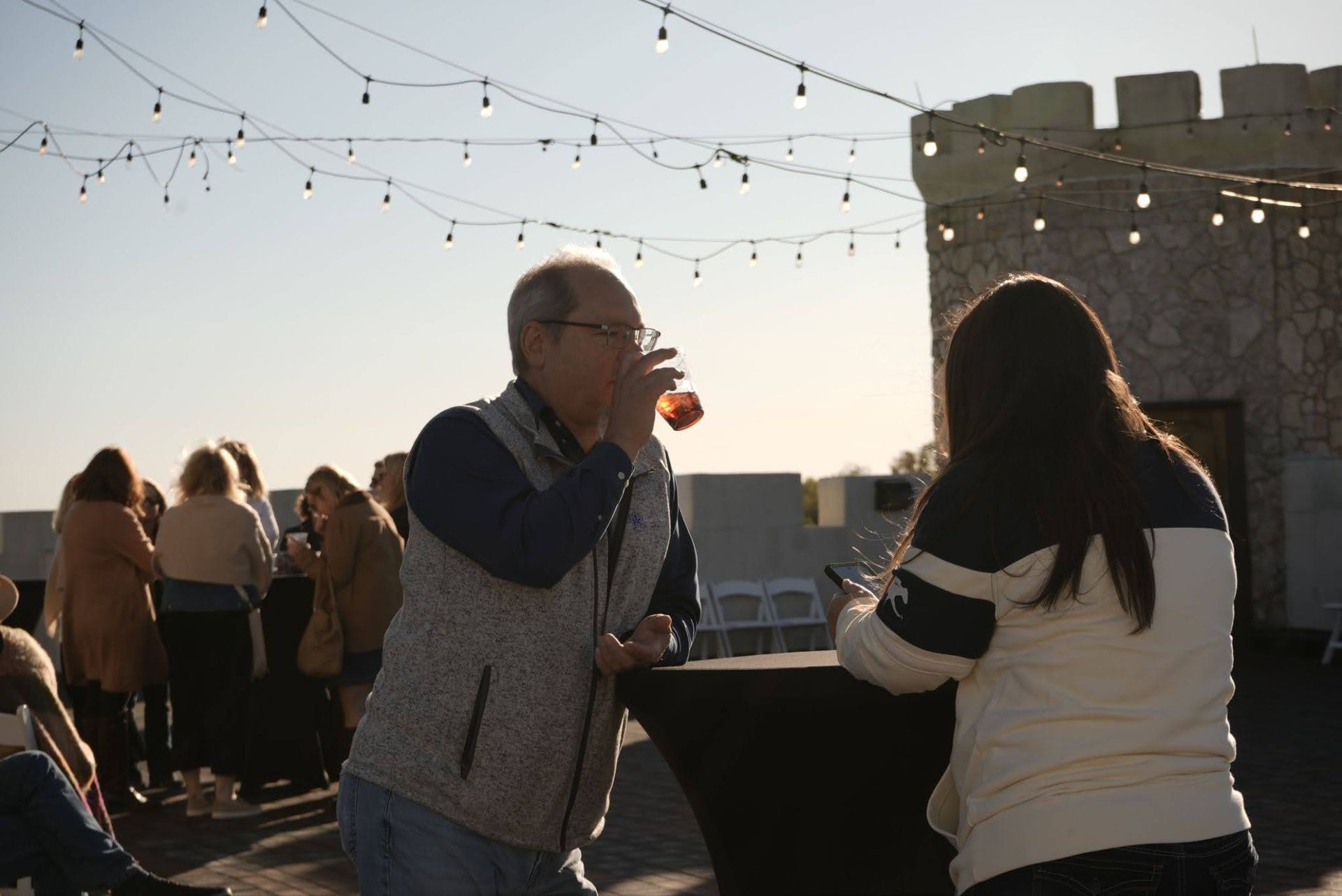 Two people chatting and drinking at an outdoor rooftop party at sunset, with string lights overhead.