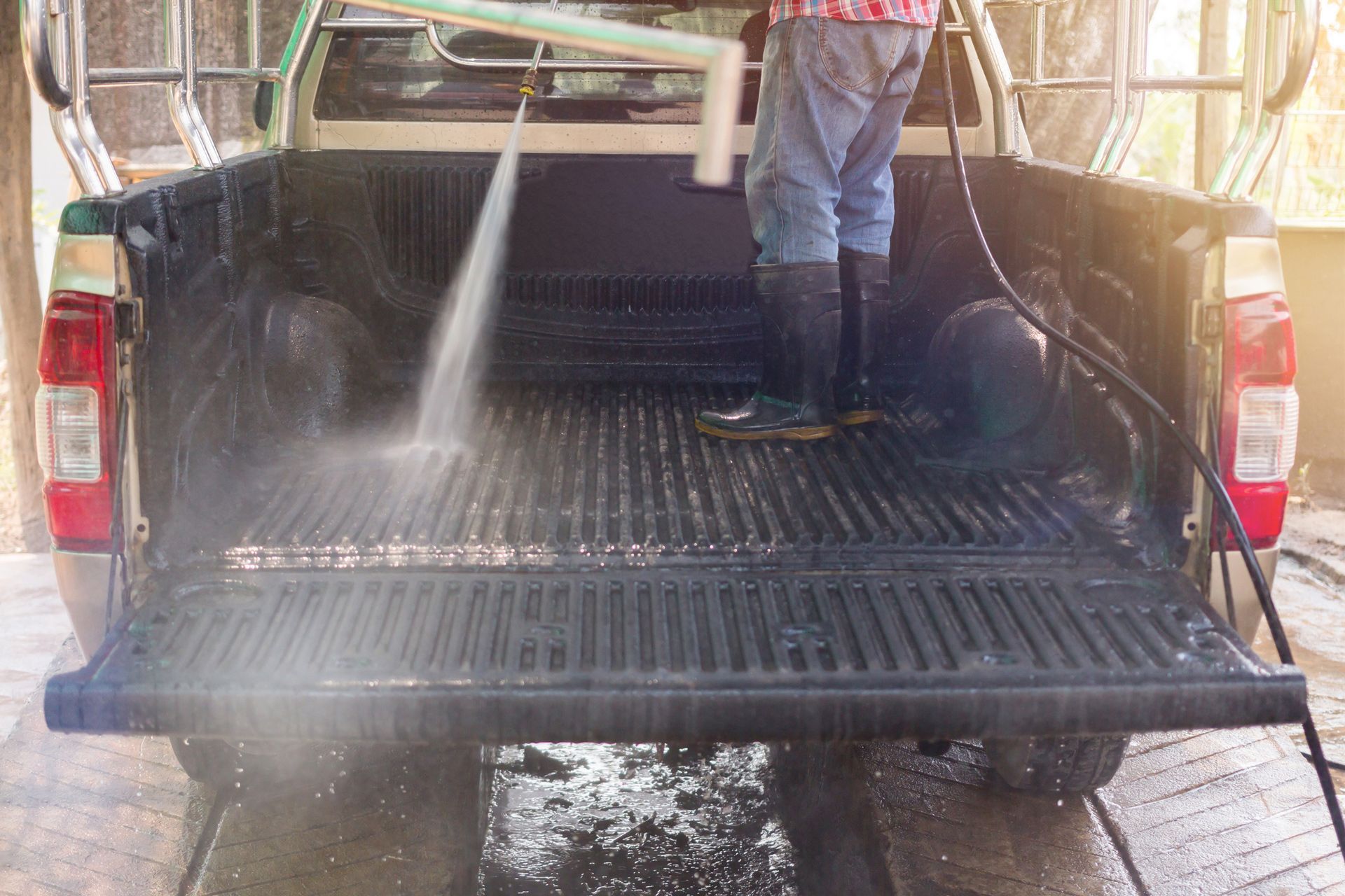 Person spraying the bed of a pickup truck with water. Dark bed with ridged liner. Outside, sunny.