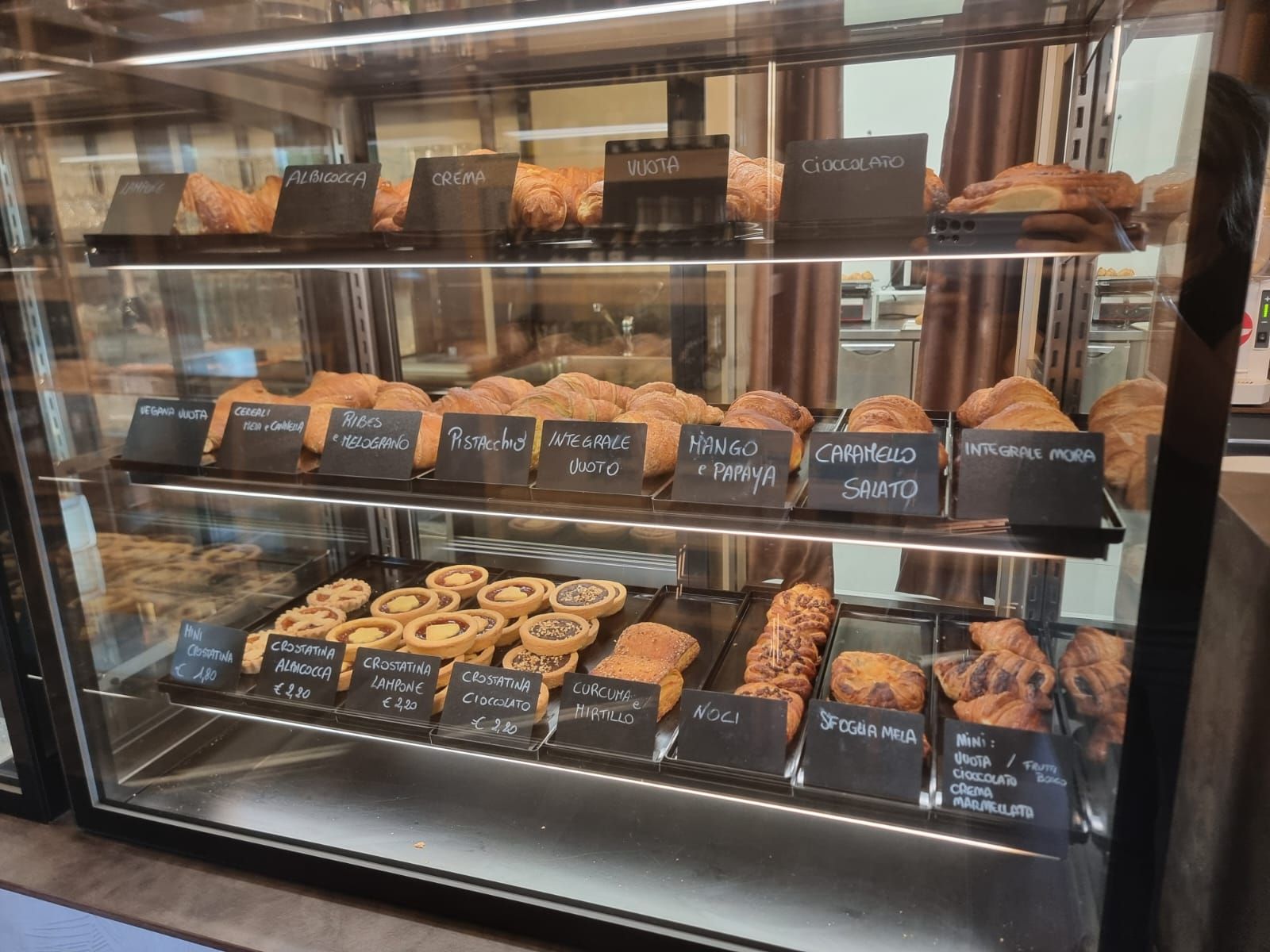 Pasticcini esposti in una vetrina da panetteria con etichette scritte a mano; vari tipi di pane.