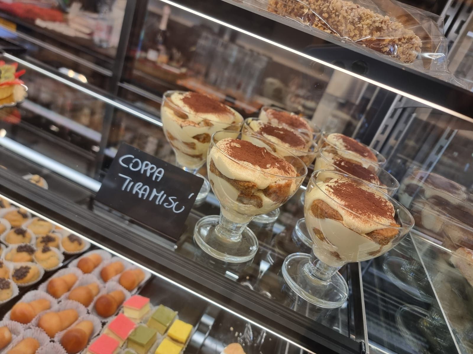 Tiramisù in coppette di vetro esposte in una vetrina da pasticceria insieme ad altri dolci.