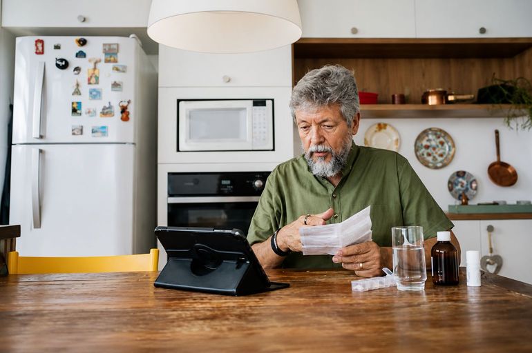 Man in a green shirt reading papers at a kitchen table with a tablet and a glass of water.