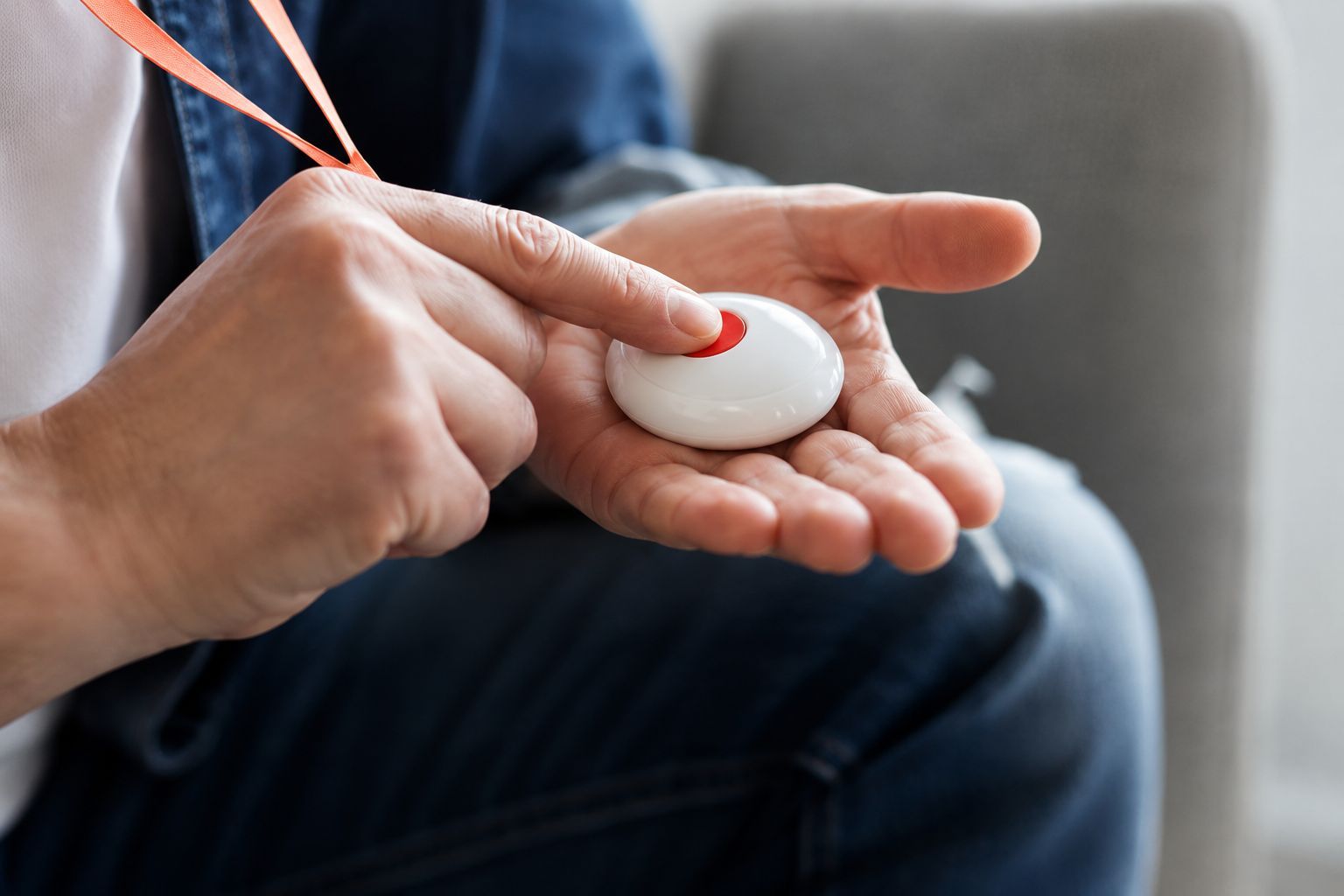 Hands holding a white medical alert device on a string, with a red button, seated on a couch