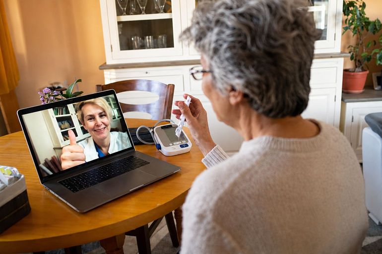 Person on a video call on a laptop at a kitchen table, holding a mug and facing the screen.