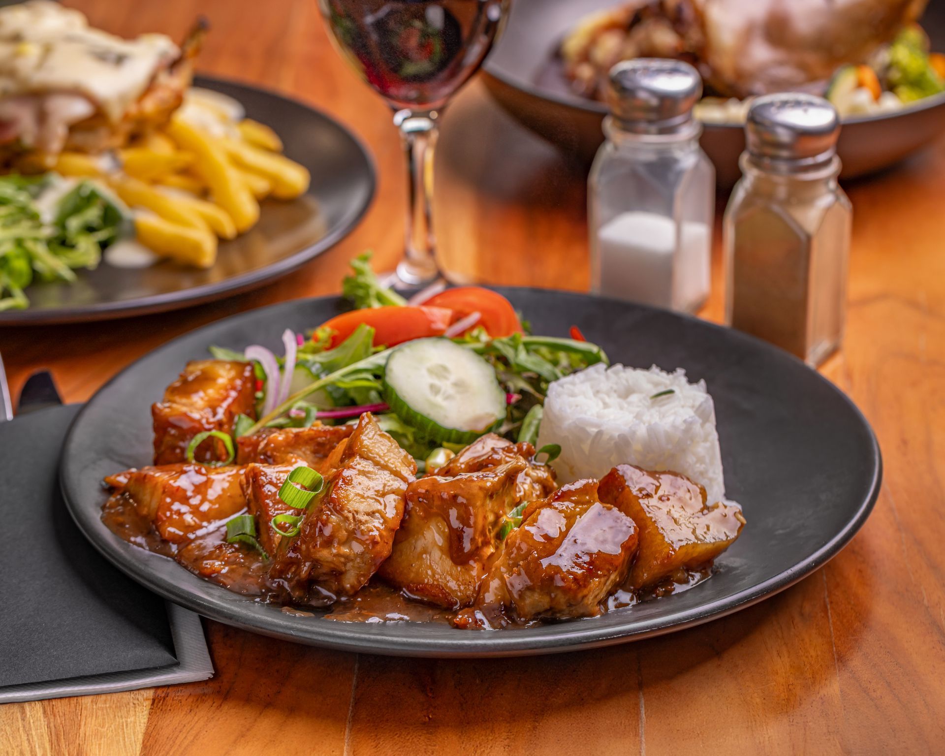 Pork dish with rice, salad, and sauce on a dark plate. Red wine and other dishes in background. — Inverell Sporties in Inverell, NSW