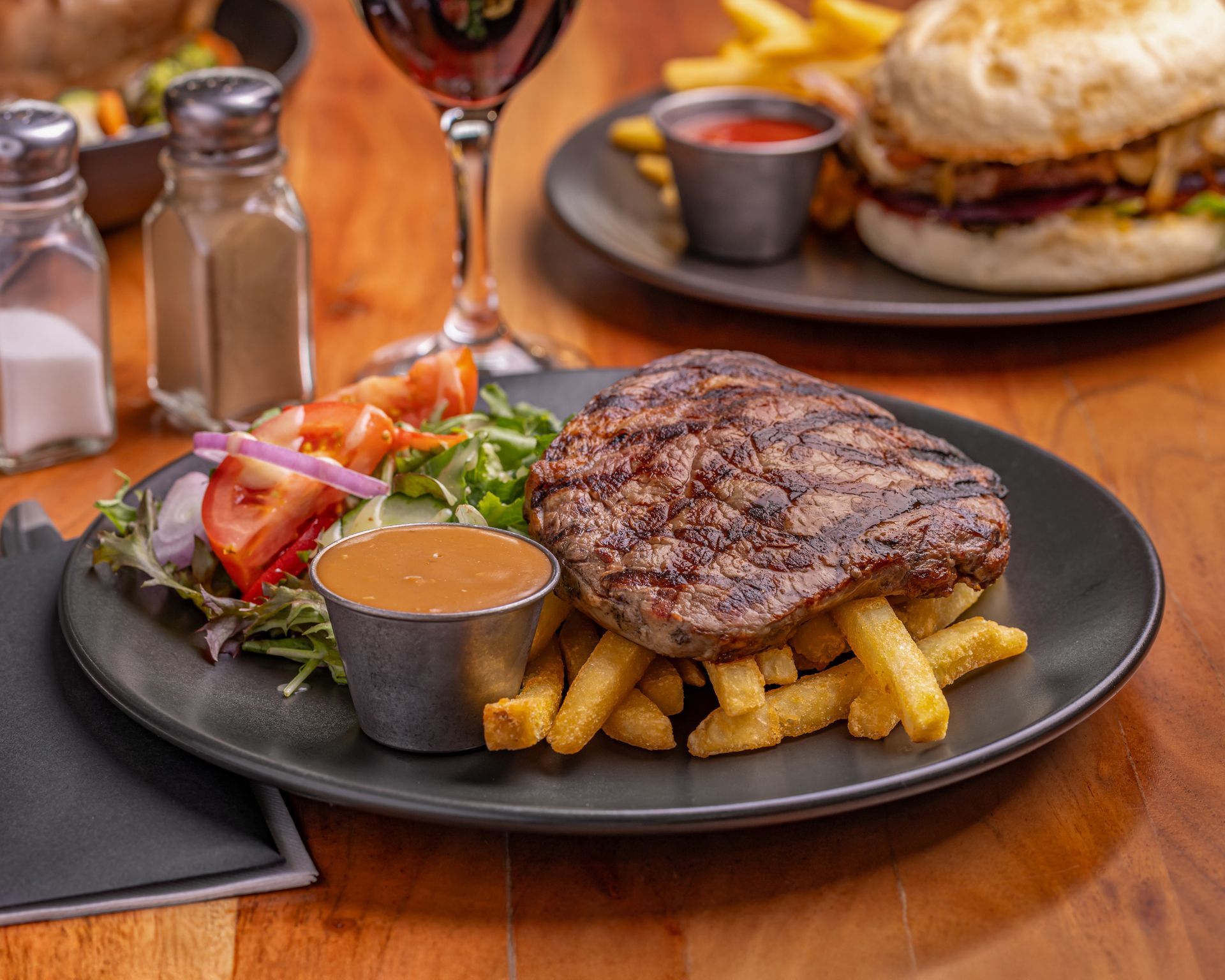 Steak with fries and gravy on a black plate, salad, wine glass, and burger in the background. — Inverell Sporties in Inverell, NSW