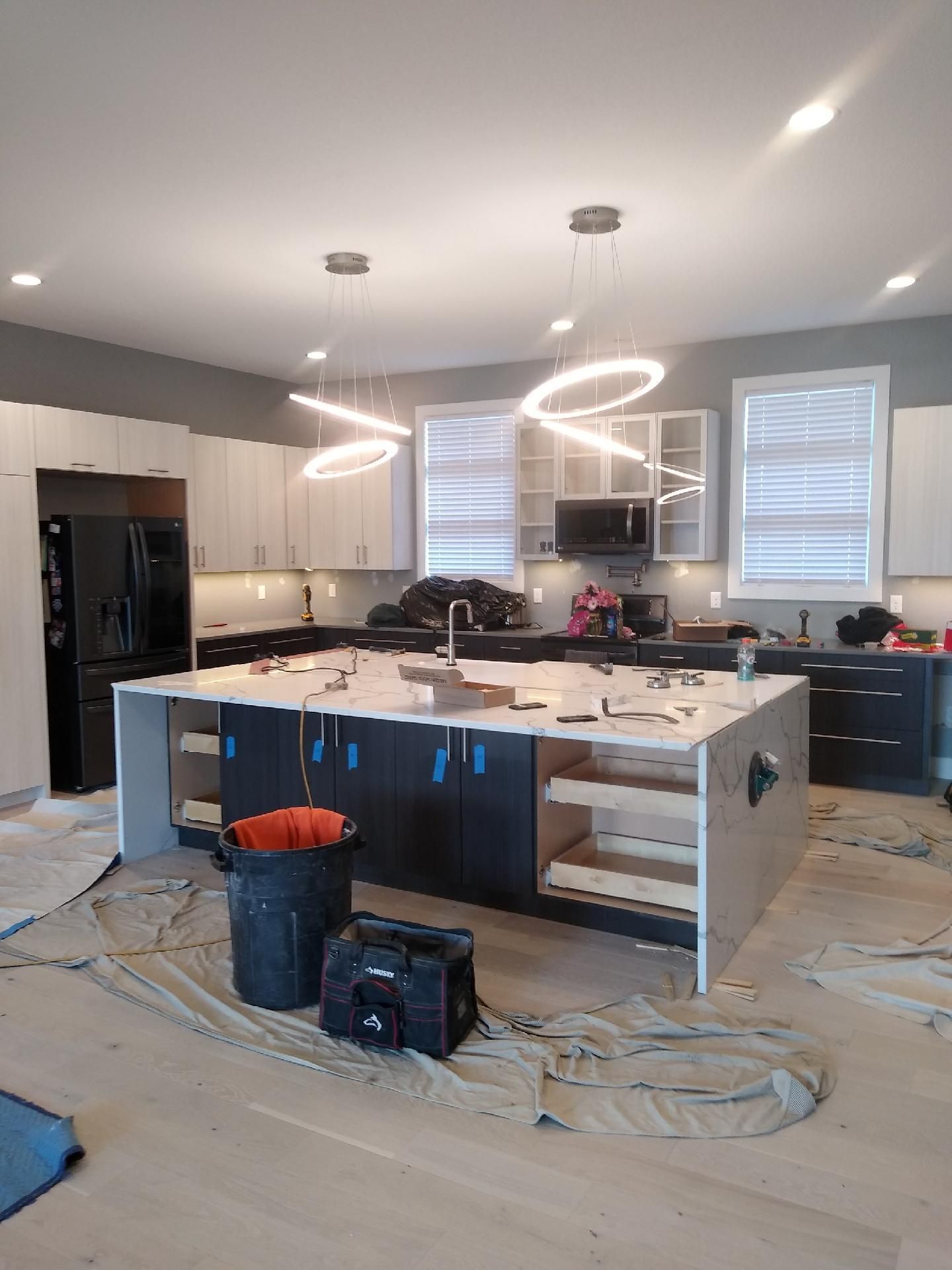 Kitchen under renovation with white cabinets, black fridge, island, and hanging pendant lights