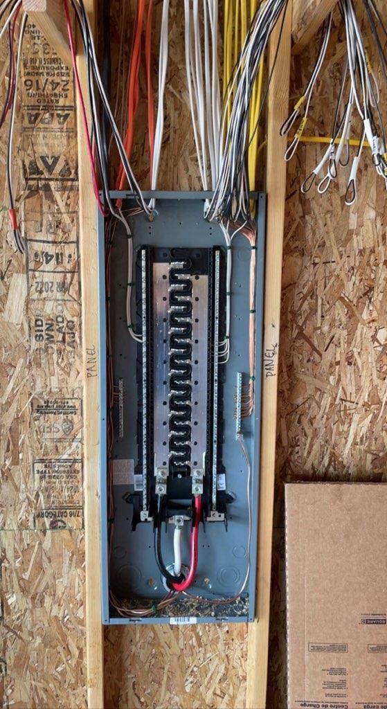 Open electrical panel with exposed wiring in a plywood-framed wall