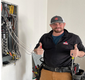 Electrician giving two thumbs up beside an open electrical panel with exposed wires.