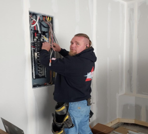 Electrician wiring an open breaker panel in an unfinished room, wearing a black hoodie and tool belt.