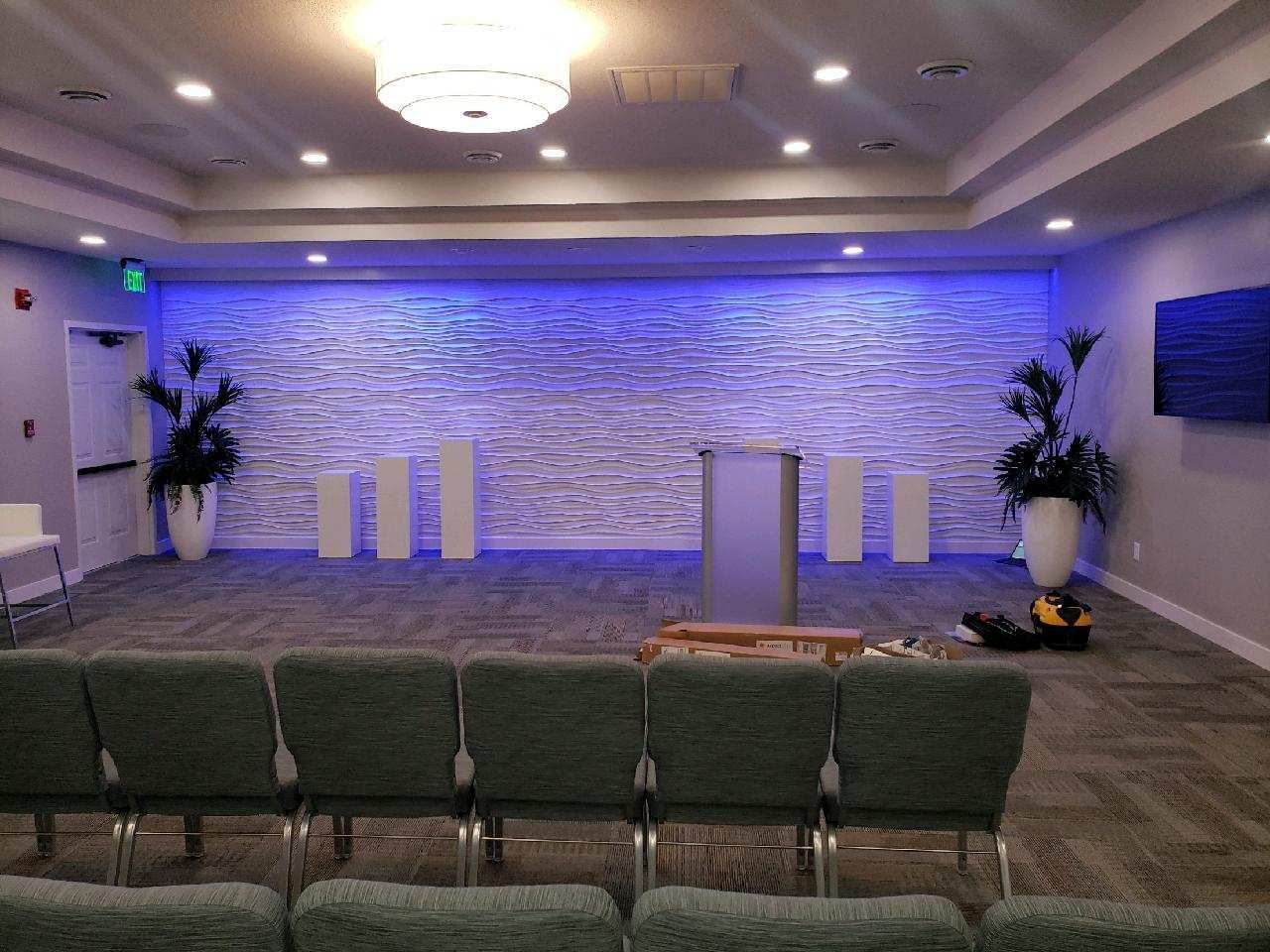 Empty church stage with stone backdrop, blue lighting, lectern, floral arrangements, and rows of green chairs