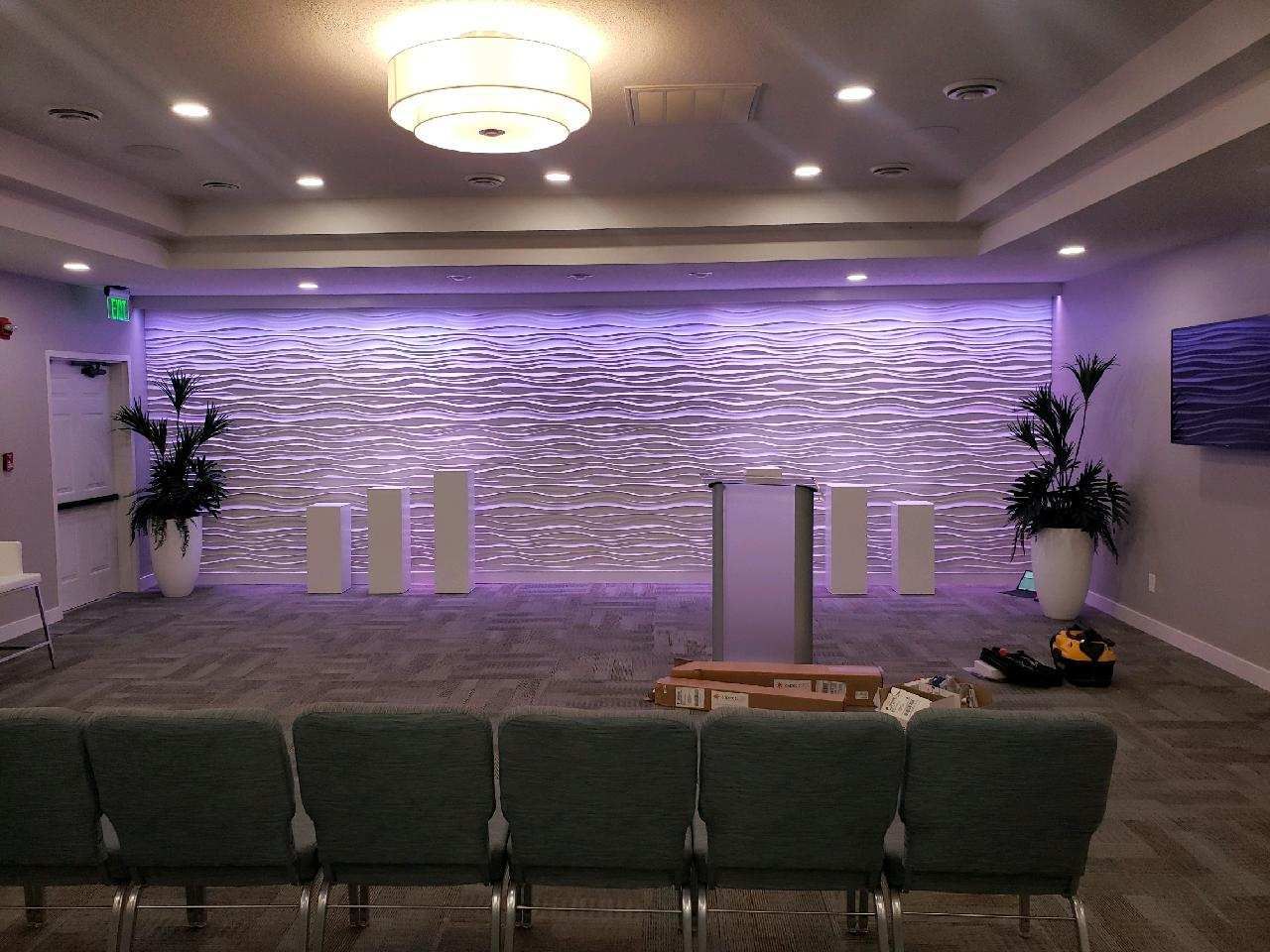 Empty event stage with purple-lit stone wall, podium, flower arrangements, and rows of chairs in front.