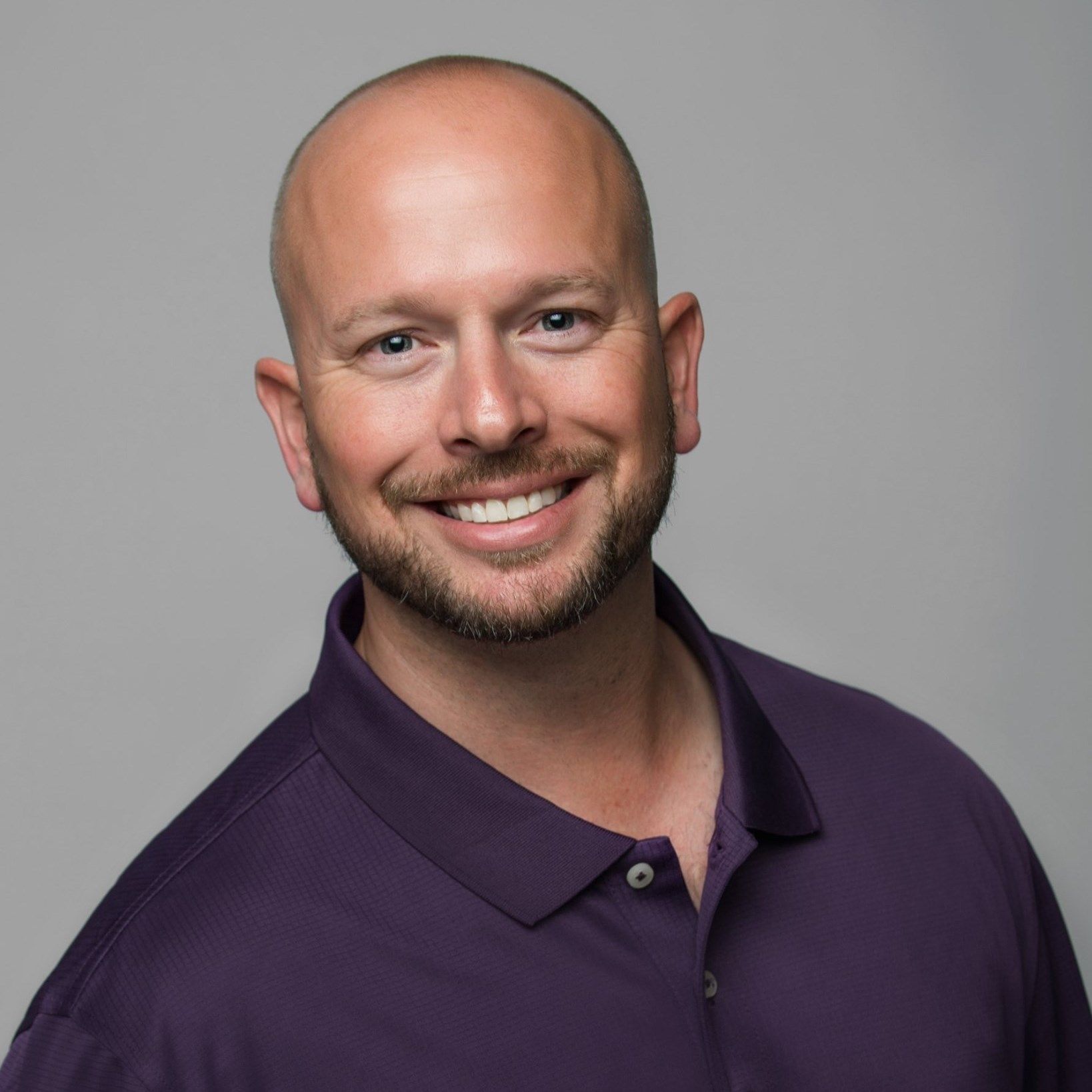 A bald man with a beard is wearing a purple polo shirt and smiling.