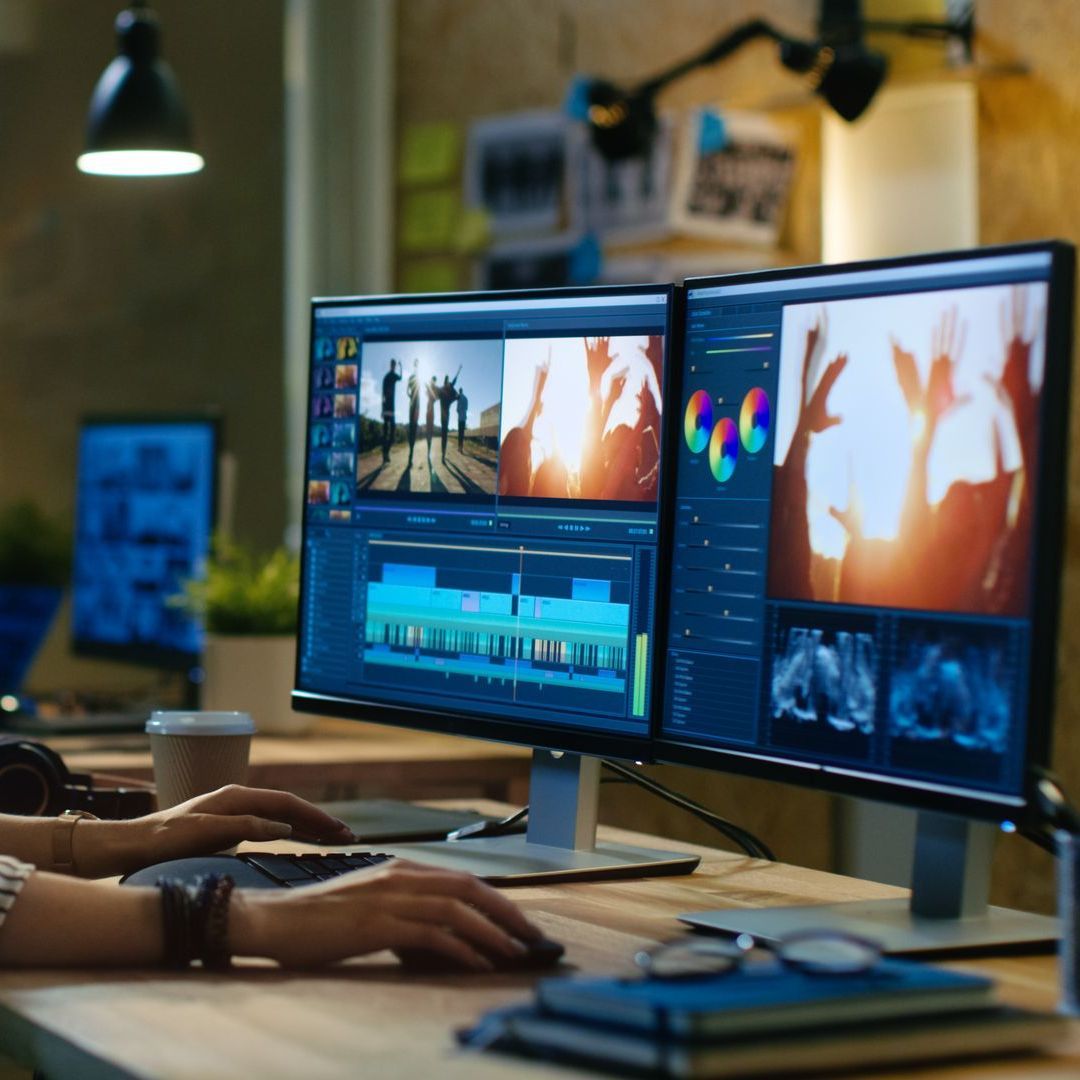 Person edits video on dual monitors in office. Sunlight, film strip, and color grading shown.