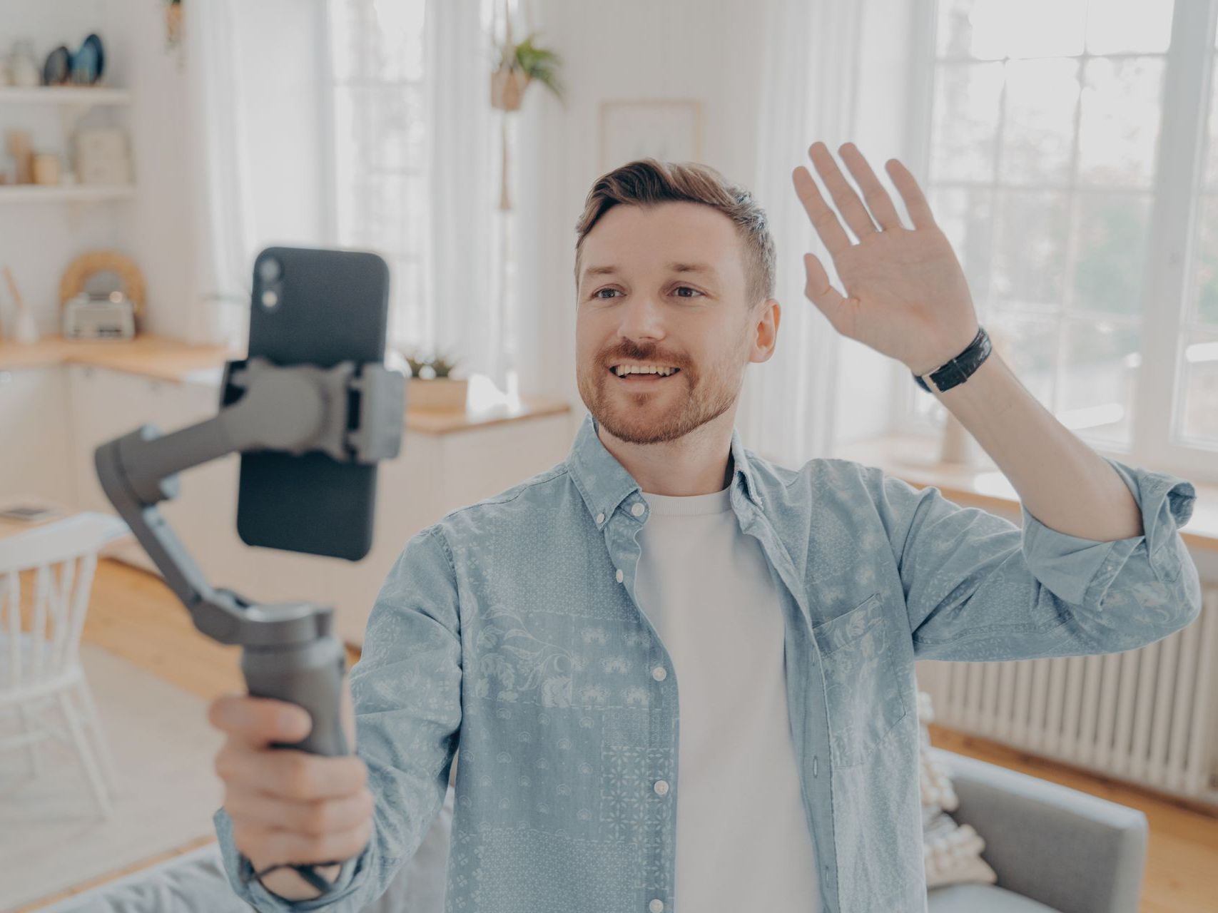 Man waves at phone on a gimbal, recording in a bright living room.