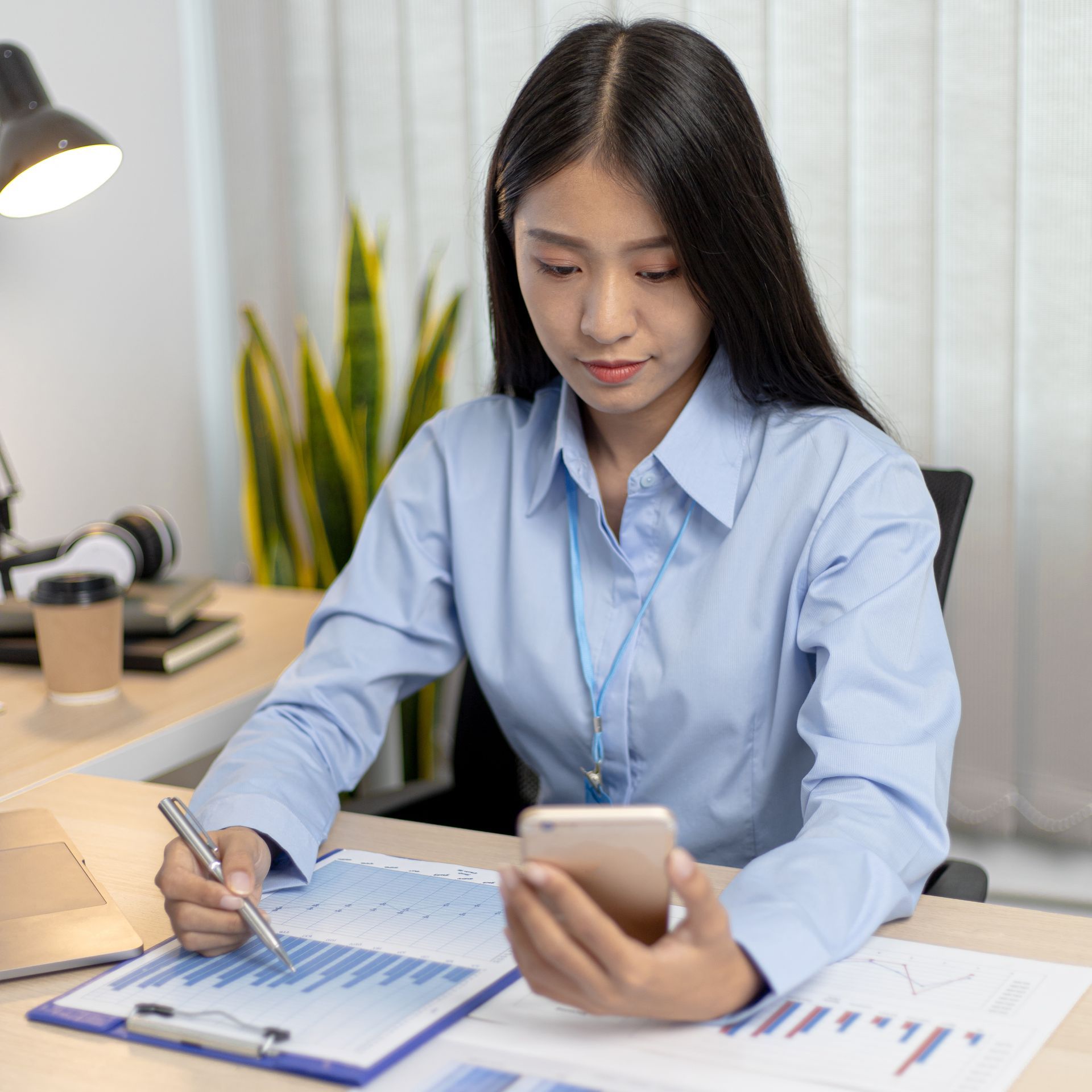 A woman is sitting at a desk using a cell phone and writing on a clipboard.