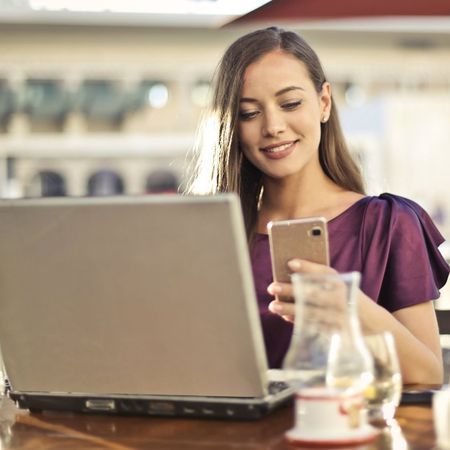 A woman is sitting at a table with a laptop and a cell phone.