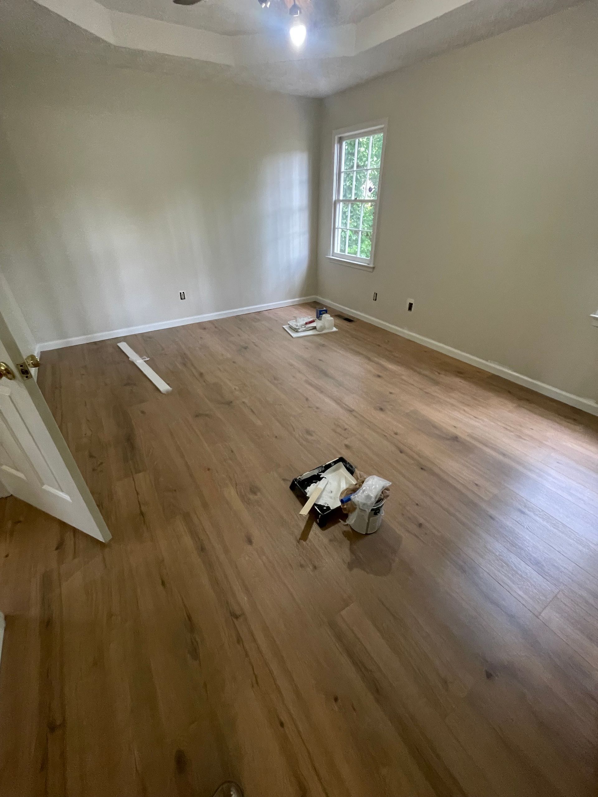 Empty room with hardwood floors, beige walls, and a small window. Paint supplies are on the floor. Empty room with hardwood floors, beige walls, and a small window. Paint supplies are on the floor.