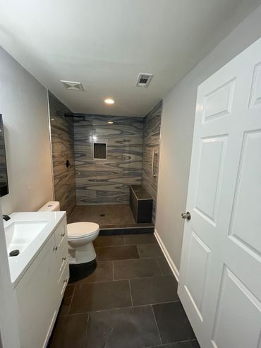 Bathroom with a gray and blue tiled shower, a white vanity and toilet, and a dark tile floor. Bathroom with a gray and blue tiled shower, a white vanity and toilet, and a dark tile floor.