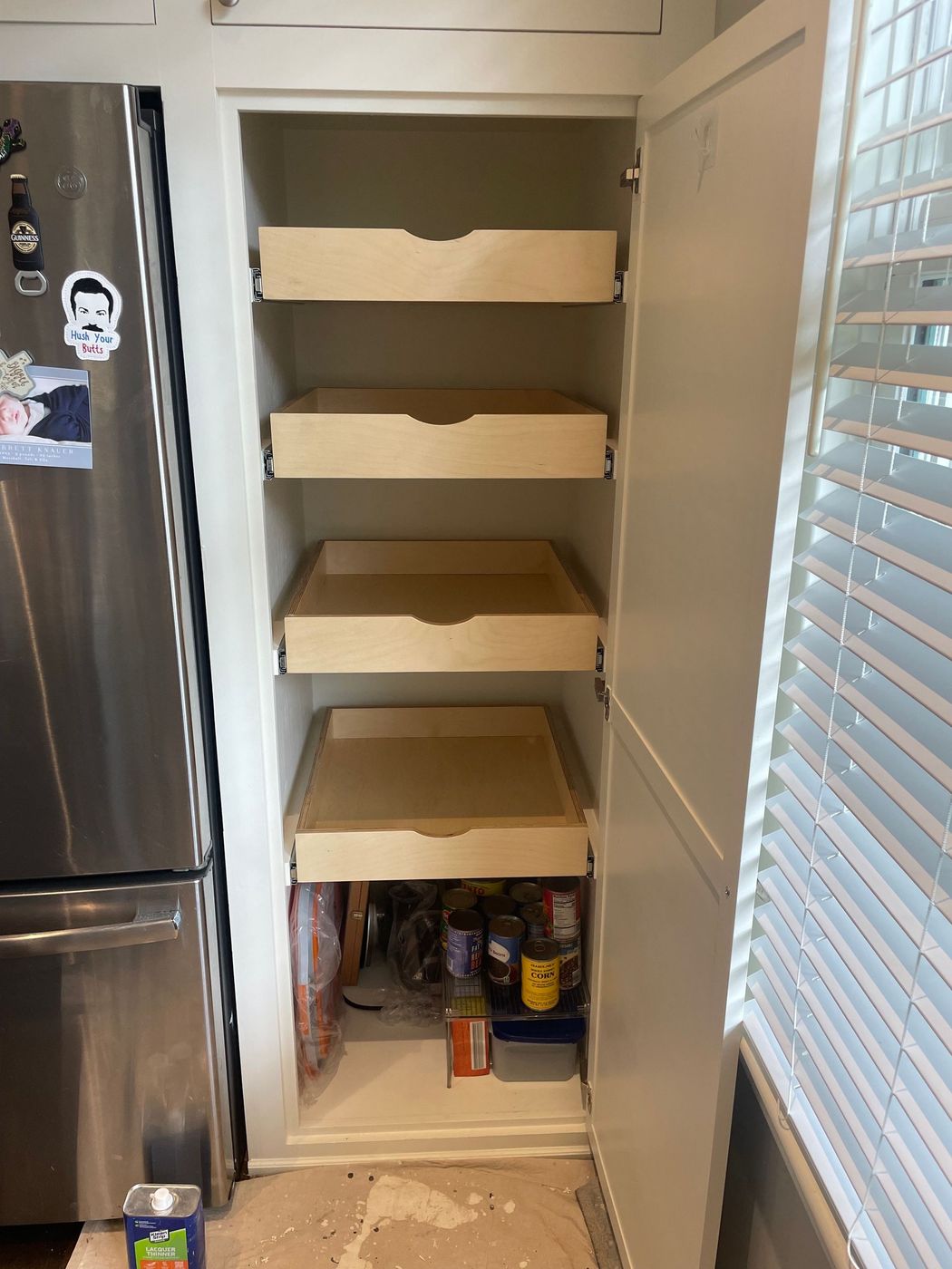 Pantry cabinet with pull-out drawers. Drawers are mostly empty. Lower shelf with food items. Stainless steel refrigerator visible. Pantry cabinet with pull-out drawers. Drawers are mostly empty. Lower shelf with food items. Stainless steel refrigerator visible.
