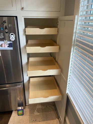 Wooden pull-out shelves inside a kitchen cabinet, next to a stainless steel refrigerator and window with blinds. Wooden pull-out shelves inside a kitchen cabinet, next to a stainless steel refrigerator and window with blinds.