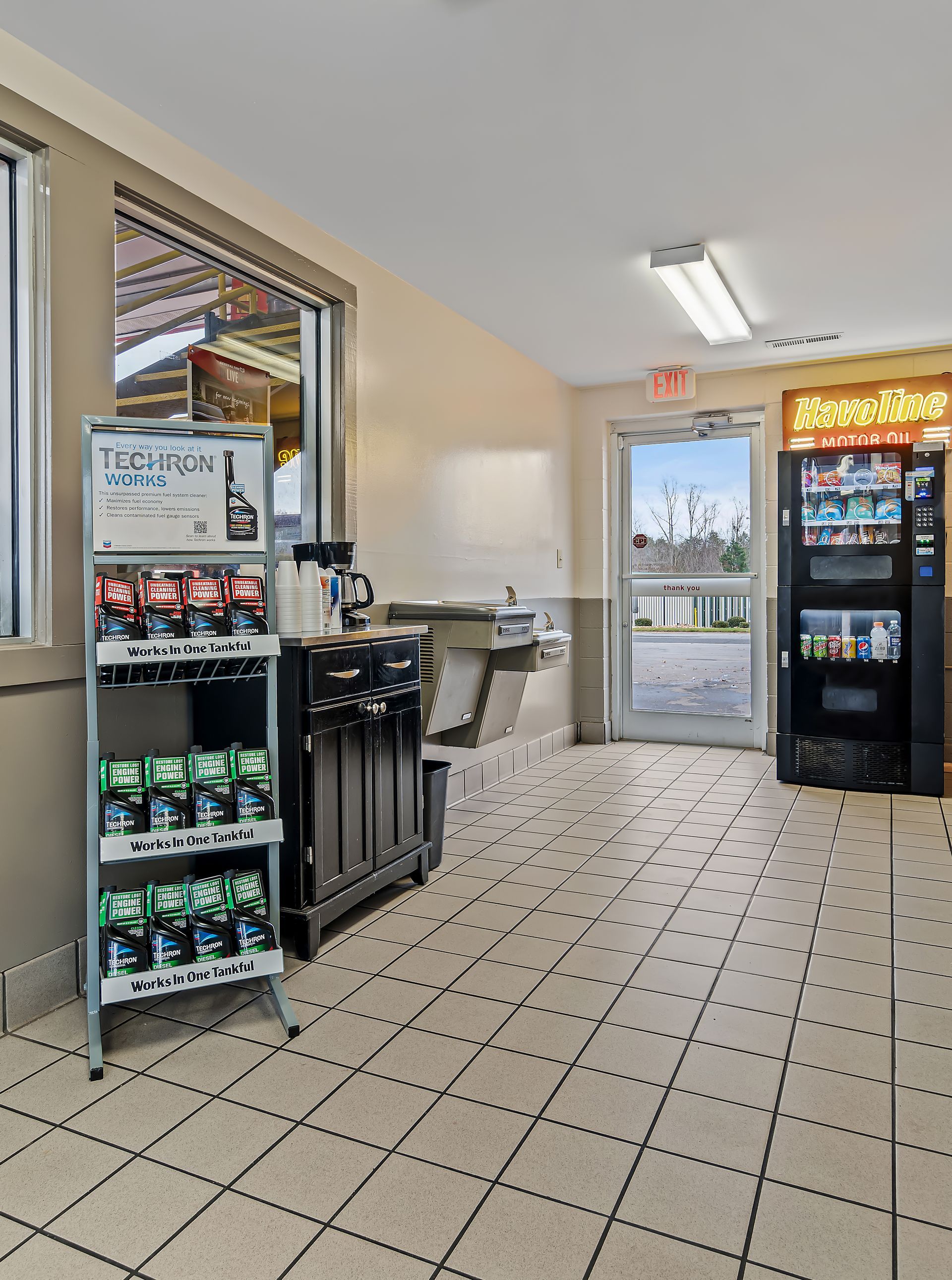 Small convenience store interior with snack racks, drink cooler, tiled floor, and glass entrance door.