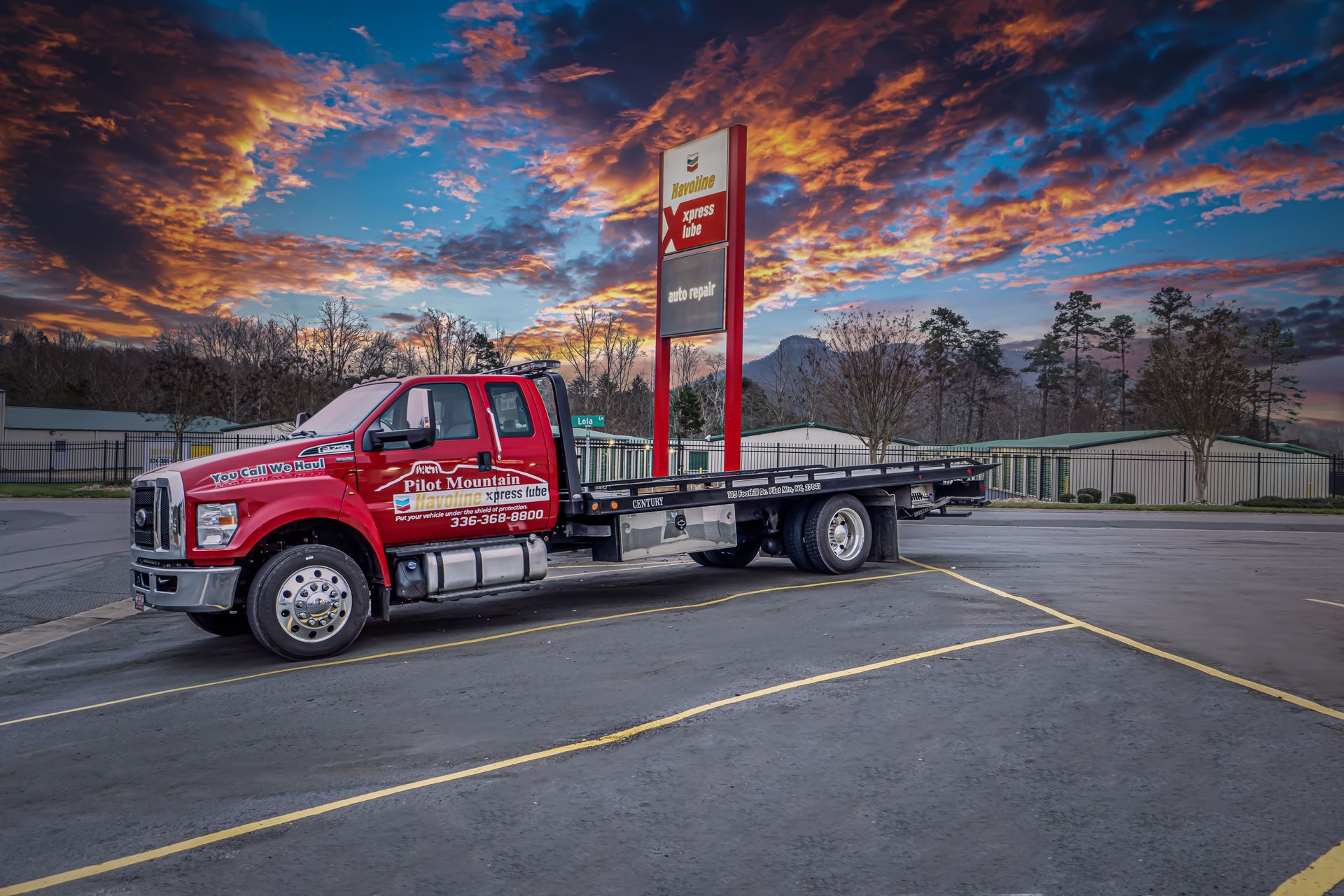 Red flatbed tow truck parked on a lot at sunset beneath a colorful sky