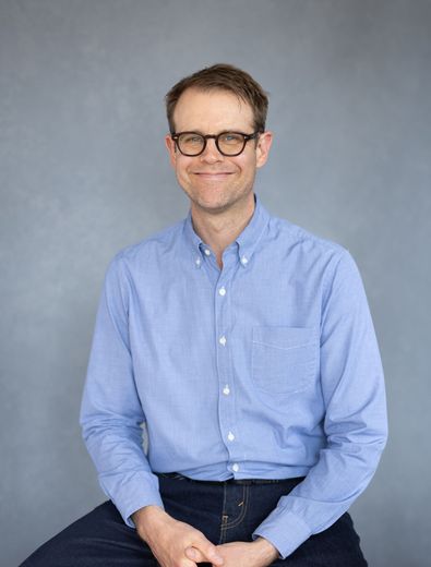 Smiling person in glasses wearing a light blue shirt against a gray backdrop.