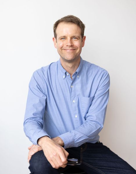 Man in a light blue shirt sitting and smiling against a plain white background