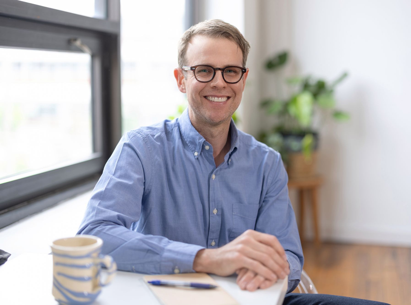 Smiling man in a blue shirt sitting at a table with a coffee mug in a bright room