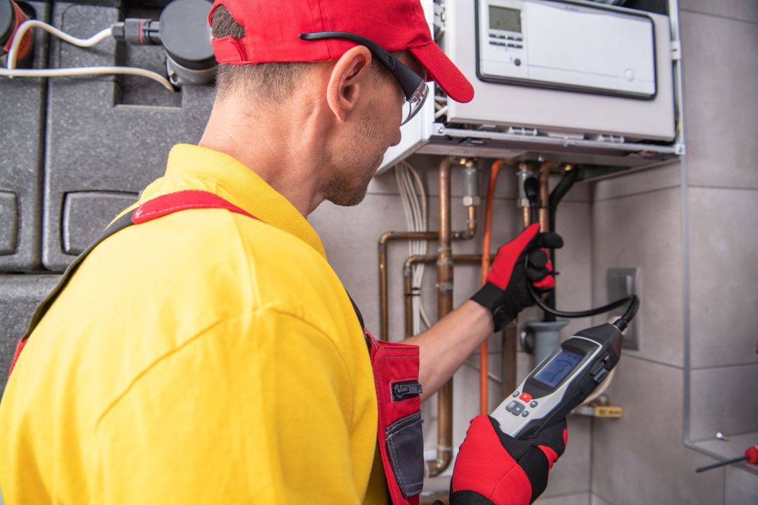 Technician in red cap and yellow shirt inspecting boiler pipes with a thermal meter in a utility room