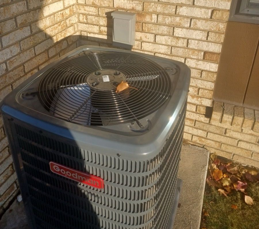 Outdoor HVAC unit beside a brick wall, with a fan grille and fallen leaves in Southeast Michigan.