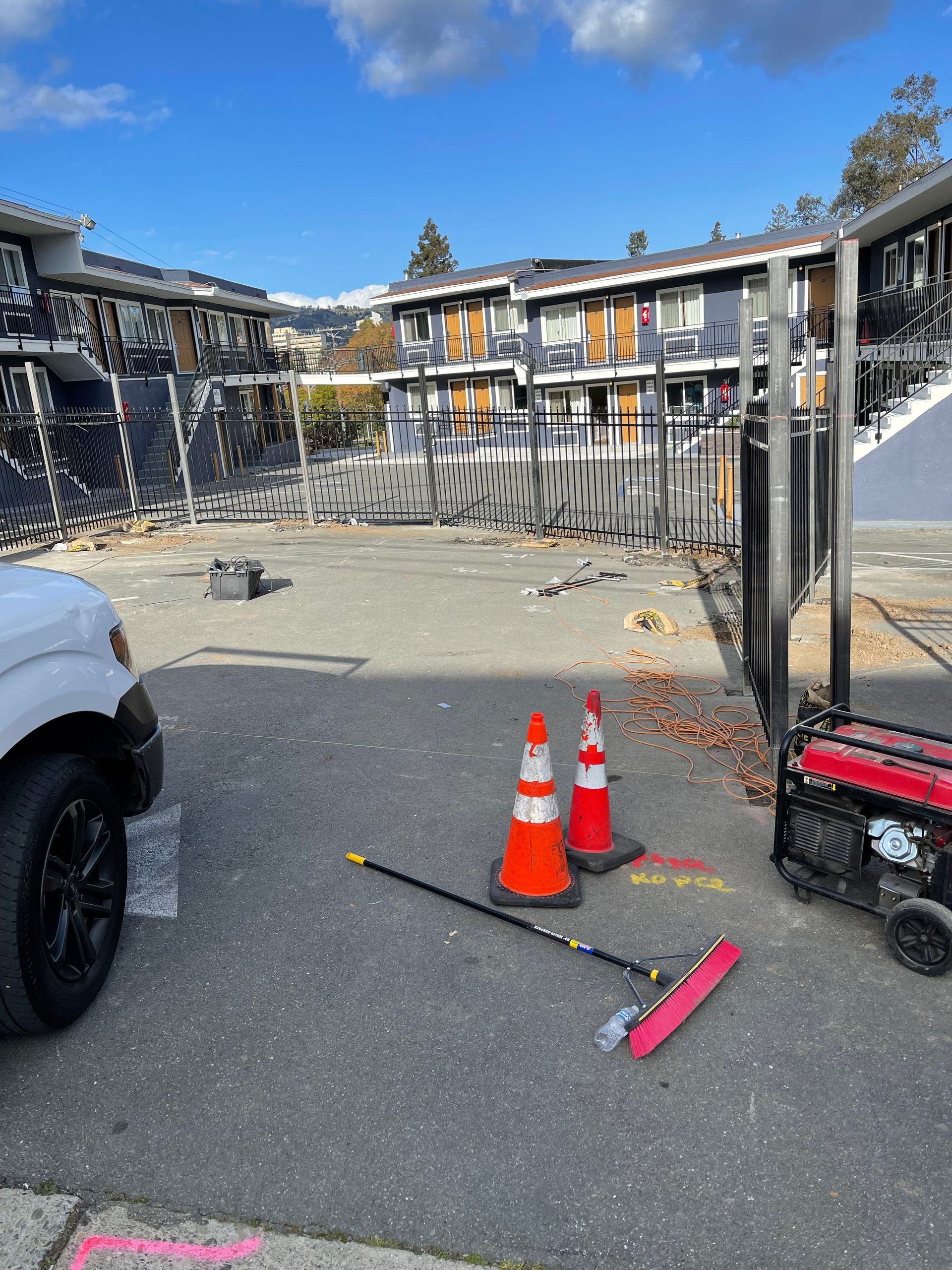 A sunny parking lot in front of a damaged apartment building, with orange safety cones, a red squeegee, and a generator.