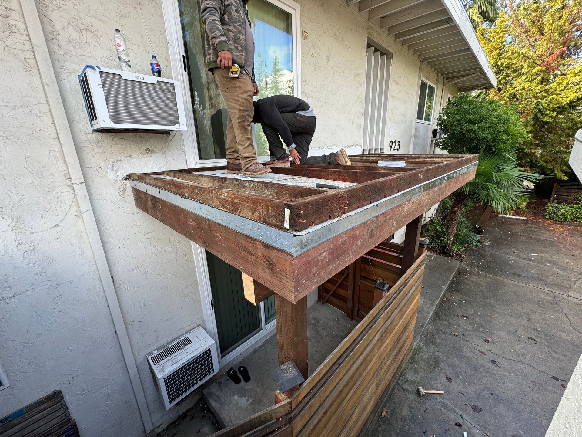 Two people work on the wooden framing of a patio roof attached to the side of a stucco house.